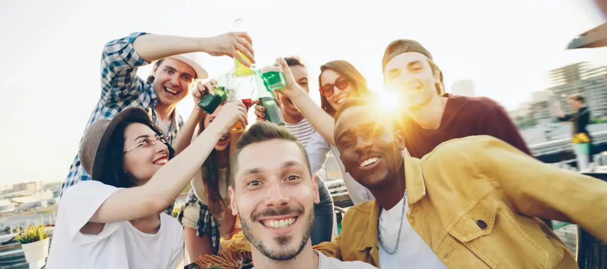 Friends on a rooftop at sunset, clinking bottles and laughing towards the camera
